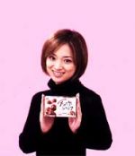 Ayu smiling, wearing a black turtleneck sweater, standing against a bright bubblegum-pink background and holding up a box of the product.